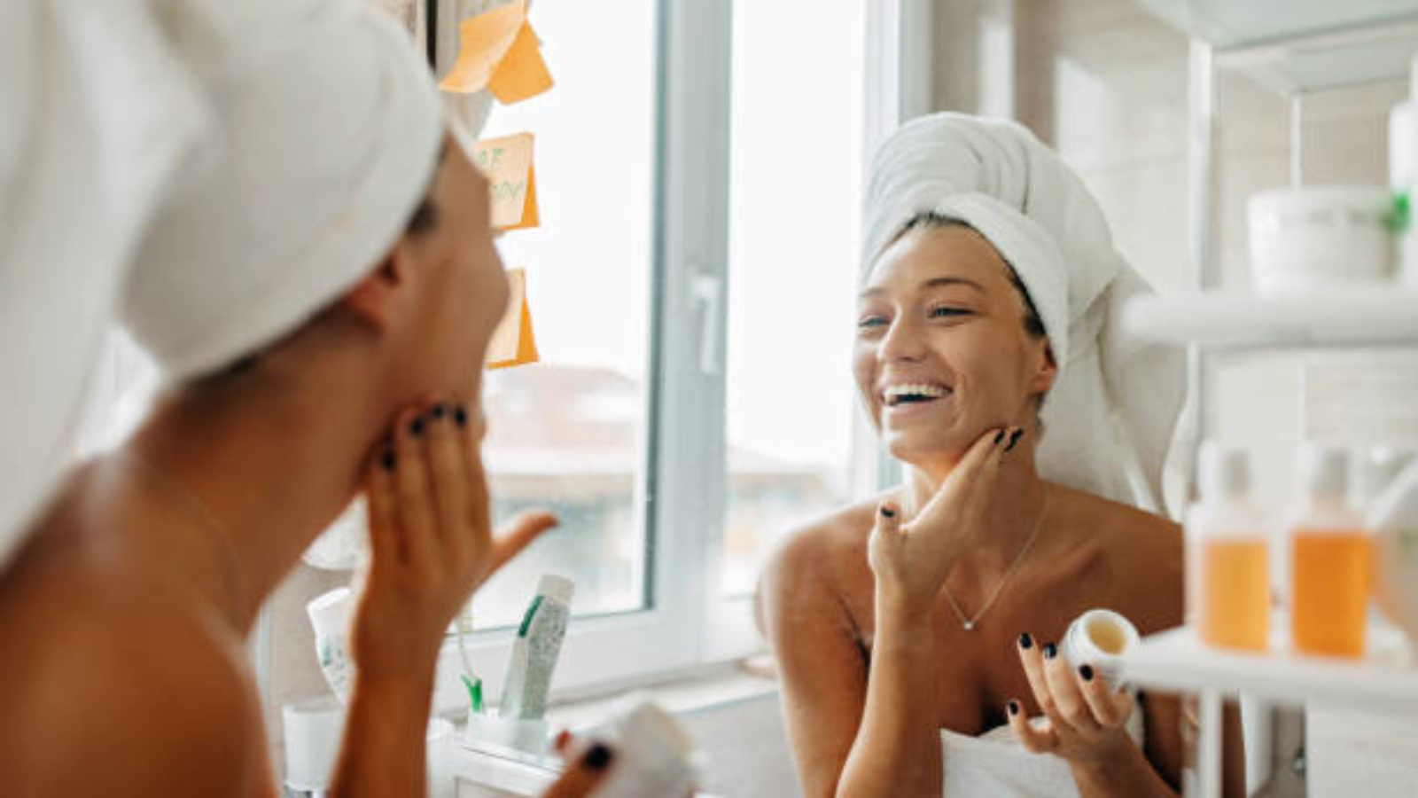 A beautiful young Caucasian woman is looking at the mirror with a big smile, while putting on her skin care products.